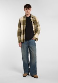 Young man wearing olive and beige plaid shirt over black tee, loose blue jeans, and tan shoes, standing against plain light background.