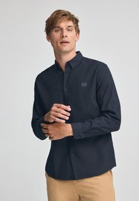 Camicia button-up a maniche lunghe di colore blu scuro, con una finitura texture, caratterizzata da un patch con logo cucito sul petto e polsini con bottone.