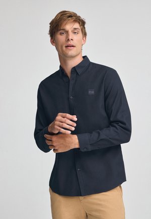 Camicia button-up a maniche lunghe di colore blu scuro, con una finitura texture, caratterizzata da un patch con logo cucito sul petto e polsini con bottone.