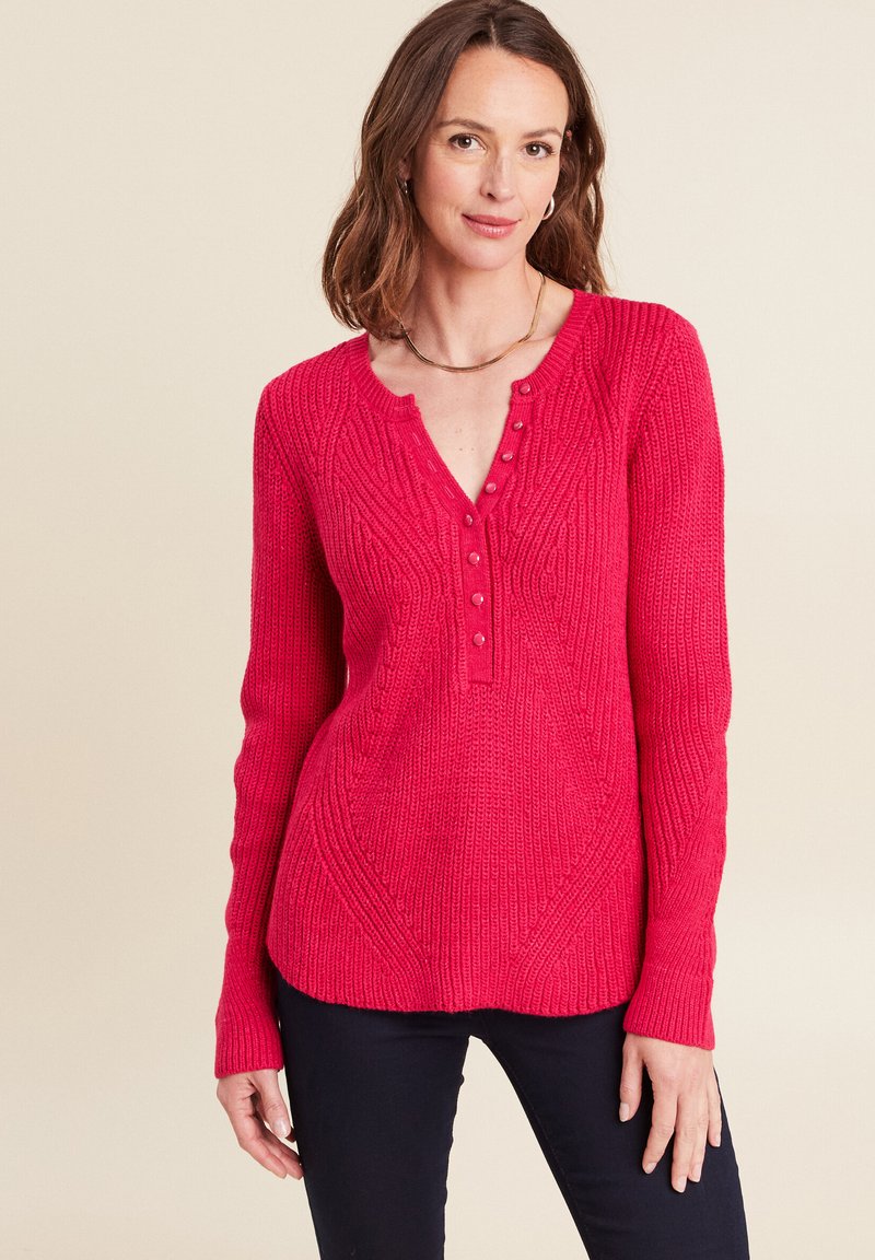Helles pinkes Rippensweatshirt mit einem V-Ausschnitt, dekorativen Knöpfen und strukturiertem Muster. Lange Ärmel verjüngen sich an den Handgelenken. Kombiniert mit dunklen Hosen.