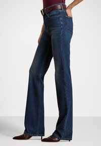 Γυναίκα που φοράει σκούρα μπλε τζιν παντελόνια με φαρδιά μπατζάκια, καφέ ζώνη και καφέ μυτερά τακούνια, στέκεται με το ένα χέρι στην τσέπη.