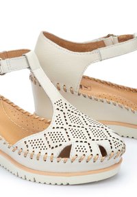 Sandali in pelle beige chiaro con tacco a zeppa, caratterizzati da motivi intagliati, dettagli di cucitura e una fascia regolabile. Suola in legno visibile.