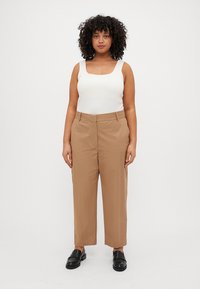 Lett hvit tanktopp med firkantet hals, parret med beige skreddersydde bukser. Modellen har på seg svarte slip-on sko og står rett opp.