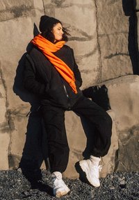 Sort puffjakke over sorte bukser, parret med et klart orange tørklæde og hvide sneakers. Baggrunden har strukturerede klippede overflader.