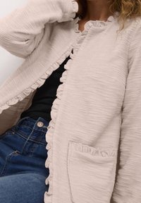 Jasnobeżowy kardigan wykonany z teksturowanej tkaniny, ozdobiony falbanami wzdłuż krawędzi i kieszeni. Charakteryzuje się luźnym krojem i otwartym przodem.