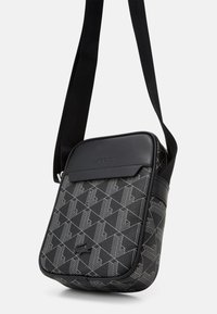 Black crossbody bag with geometric pattern in gray. Features a smooth black leather flap and adjustable strap. Zipper closure at the top.