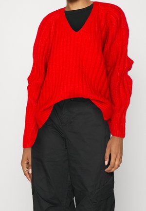 Maglione rosso a coste con scollatura a V, maniche lunghe e vestibilità rilassata. Indossato sopra una maglietta nera, abbinato a pantaloni cargo neri.