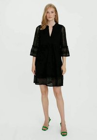 Vestido preto de renda com decote em V, mangas flare de três quartos e silhueta ampla. Apresenta recortes em padrão floral e barra até ao joelho.