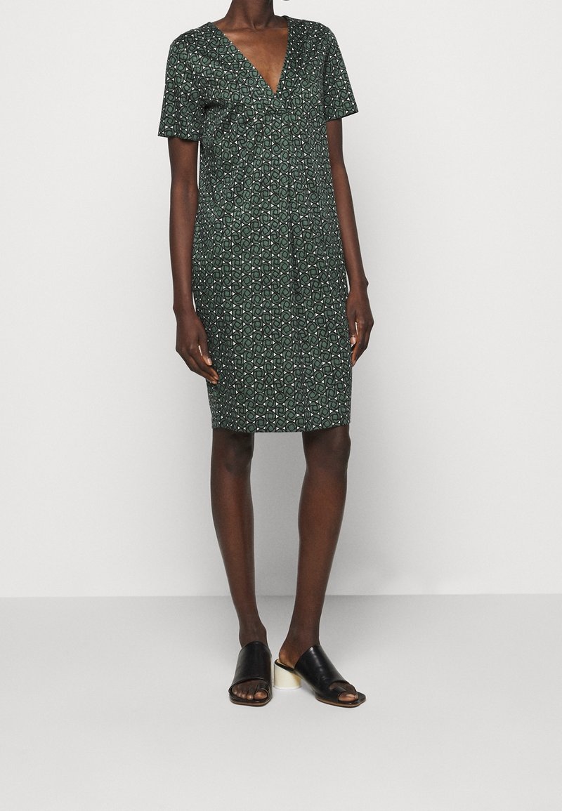 Femme portant une robe vert foncé longueur genoux avec motif géométrique, manches courtes, encolure en V et sandales à talons noires à enfiler.