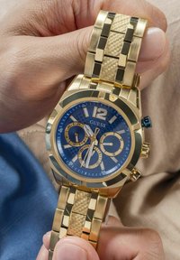 Auksinės spalvos chronografo laikrodis su mėlynu ciferblatu, tekstūruotu paviršiumi ir datos rodmeniu. Auksinė ir raštuota apyrankė, švytinčios žymekliai ir apvalus korpusas.