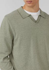 Lichtgroene, gestructureerde gebreide trui met V-hals en puntige kraag. Heeft dunne, horizontale strepen en lange mouwen, gedragen over een wit overhemd.