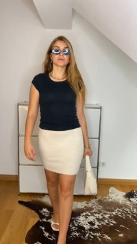 Maglietta nera aderente, gonna beige a vita alta, borsa bianca e sandali bianchi. Capelli lunghi e occhiali da sole arricchiscono l'outfit.