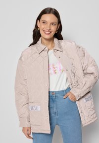 Gevoerde, gestepelde jas in lichtbeige met een kraag, drukknoopsluitingen en logopatches. Gemaakt om te dragen over een wit shirt en spijkerbroek.