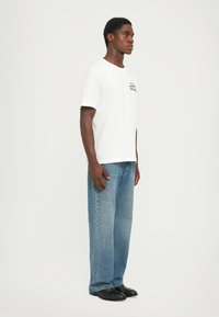 Tricou alb din bumbac cu text imprimat pe pieptul stâng, purtat cu blugi de denim ușori, cu croială largă, de culoare albastru deschis și încălțări negre.