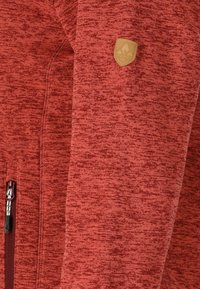 Röd fleecejacka med ett texturerat mönster, utrustad med dragkedja och en liten läderlogotyp på överarmen.