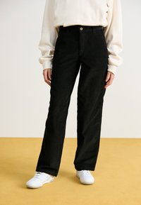 Μαύρο καμπαρντίνα παντελόνι με ίσια γραμμή, με κλείσιμο κουμπιού και πλαϊνές τσέπες, συνδυασμένο με λευκά αθλητικά παπούτσια.