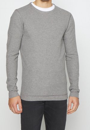 Mand iført en lys grå langærmet crewneck sweater over en hvid undertrøje og sorte bukser mod en ensfarvet baggrund.