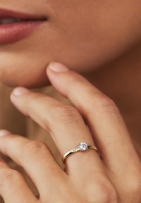 Goldring mit einem glatten Band, das einen einzelnen runden Diamant als Mittelpunkt aufweist, vor einem neutralen Hautton-Hintergrund.