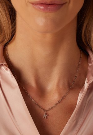 Zilveren ketting met verlengde schakels en een kleine hanger in de vorm van de letter "K." Draagt over een roze satijnen blouse.
