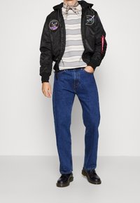 Czarna kurtka bomber z naszywkami; szary sweter w paski; niebieskie dżinsy; czarne buty; model pozujący na jednolitym tle.