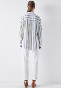 Chemise à manches longues, boutonnée, blanche avec des rayures verticales bleues, vertes et jaunes. Associée à un pantalon blanc et des sandales à talons carrés marron.