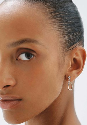 Mujer con cabello liso recogido, que lleva un pendiente dorado colgante adornado con tres pequeños diamantes redondos.