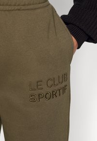 Oliwkowe dresy o miękkiej strukturze, z haftowanym logo "LE CLUB SPORTIF" w ciemniejszym odcieniu. Elastyczny pas z kieszeniami.