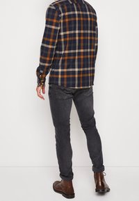 Långärmad rutigt skjorta i marinblått, orange och beige; ihop med mörkgrå jeans och bruna läderstövlar. Synliga knappmanschetter och detaljer på baksidan.