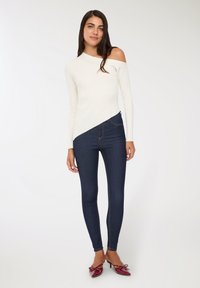 Kobieta w asymetrycznym, białym swetrze bez ramion, ciemnoniebieskich obcisłych dżinsach i bordowych szpilkach z kokardkami, stojąca na białym tle.