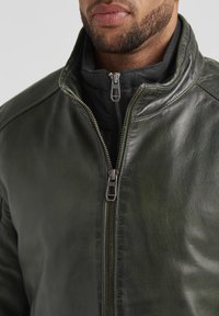 Dunkelgrüne Lederjacke mit hohem Kragen, ausgestattet mit einem silbernen Reißverschluss und einer schwarzen inneren Schicht, die eine glatte Textur und ein elegantes Design zeigt.