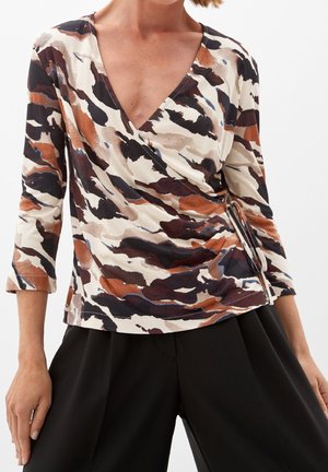 Vrouw draagt een lange mouwen wikkeltop met bruine, zwarte en beige abstracte patronen en zwarte wijde broek.