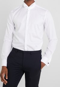Camicia bianca con colletto, realizzata in tessuto morbido, con chiusura a bottoni e maniche lunghe, abbinata a pantaloni scuri con texture.