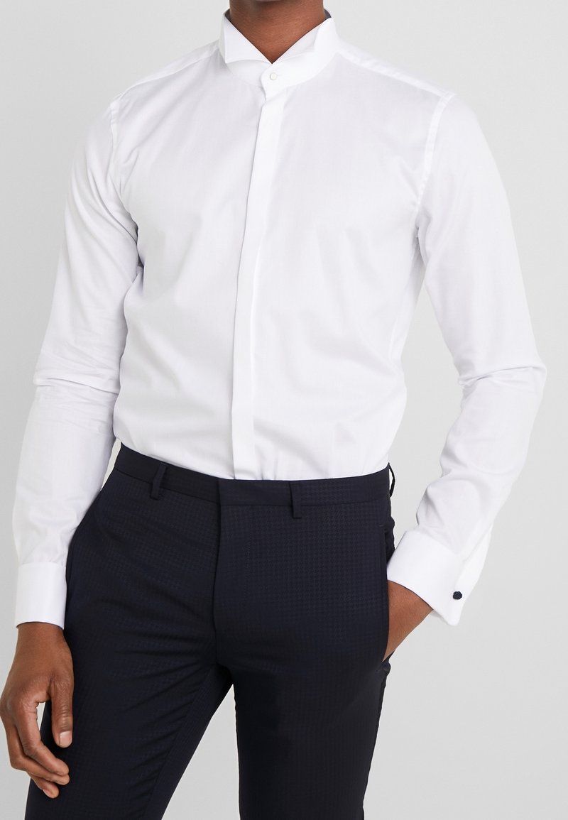 Camicia bianca con colletto, realizzata in tessuto morbido, con chiusura a bottoni e maniche lunghe, abbinata a pantaloni scuri con texture.