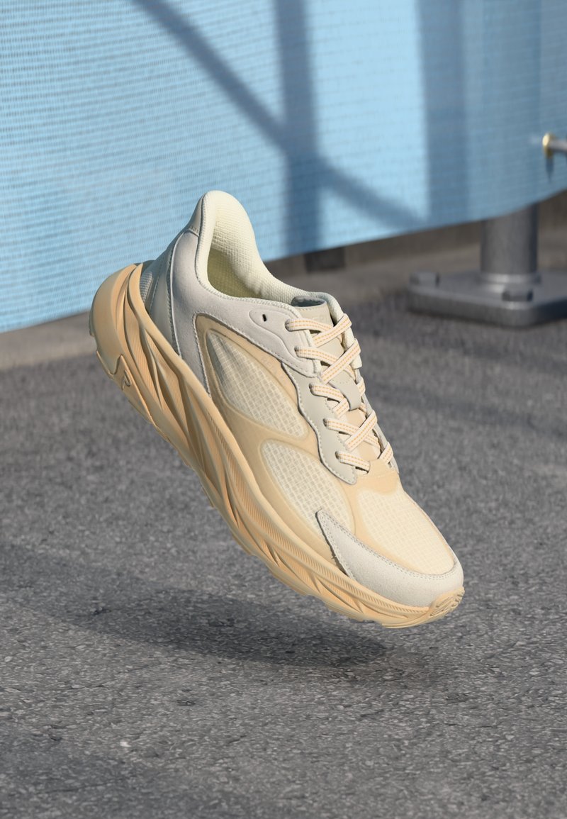 Beige sneakers med en teksturert overdel, mesh-paneler og lysfargede snøreb�ånd. De har en tykk gummisåle med en bølgelignende design.