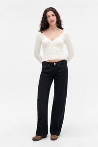 Weißes, geripptes Langarmoberteil mit Bindedetail in der Taille, kombiniert mit schwarzen, weiten Jeans mit Knopf und seitlichen Taschen.