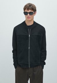 Giubbotto bomber nero con un pannello frontale in suede, maniche in maglia, chiusura frontale con zip e colletto alto. Indossato sopra una maglia scura con pantaloni marroni.