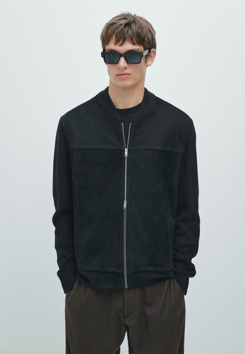 Giubbotto bomber nero con un pannello frontale in suede, maniche in maglia, chiusura frontale con zip e colletto alto. Indossato sopra una maglia scura con pantaloni marroni.