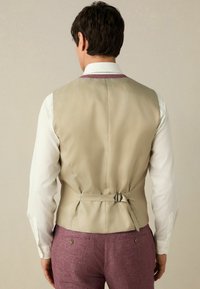 Beige vest med en glat tekstur, justerbar rygrem og lilla detaljer, parret med en hvid skjorte og lyse lilla bukser.