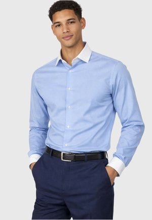 Jeune homme portant une chemise habillée bleu clair à col et poignets blancs, pantalon bleu marine et ceinture noire, les mains dans les poches, fond neutre.