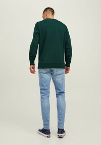 Uomo che indossa una camicia a maniche lunghe verde scuro, jeans azzurri chiari e sneakers blu navy, in piedi di spalle contro uno sfondo semplice chiaro.