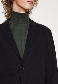 Schwarzer Blazer aus strukturiertem Stoff, mit einem einzelnen Knopf, Revers und Brusttasche, über einem grünen Rollkragenpullover getragen.