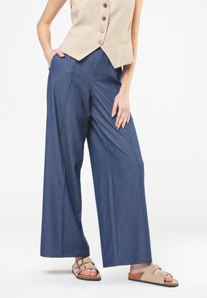 Donna con pantaloni blu a gamba larga, gilet beige senza maniche con bottoni e sandali di sughero beige con due cinghie e fibbie.