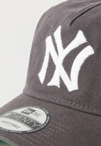 Grå kasket med hvidt broderet "NY"-logo. Har en buet skygge og justerbar strop. Stoffets tekstur er lærredslignende og blød.