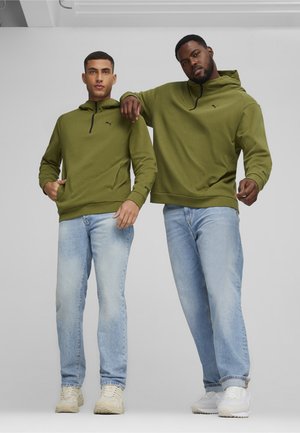 Grønne hættetrøjer med halv lynlås, parret med lyseblå jeans og beige sneakers, båret af to modeller, der poserer sammen.