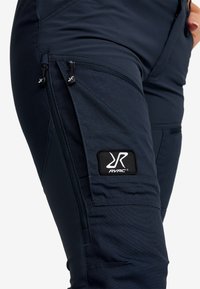 Mörkblåbyxor med textilstruktur, siddragkedjor och en svart RVRC-logotyp. Designade med praktiska detaljer för funktionalitet.