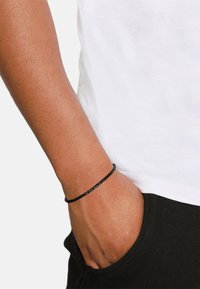 Geflochtenes schwarzes Armband aus strukturiertem Material, am Handgelenk getragen. Hand ruht in der Tasche einer schwarzen Hose, über einem weißen T-Shirt.