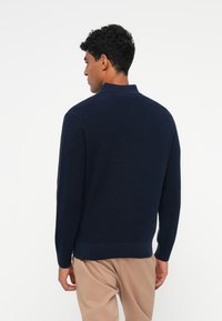Pulover tricotat de culoare bleumarin, cu guler, dintr-o țesătură texturată și margine ondulată, văzut din spate. Design simplu și finisare moale.