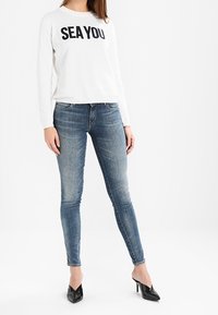 Camisa blanca de manga larga con "SEA YOU" en texto negro, combinada con jeans ajustados de color azul claro y zapatos de tacón puntiagudos negros.