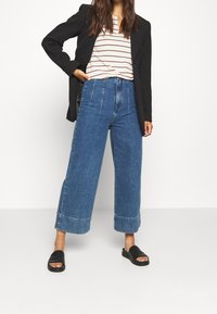 Culottes en denim bleu taille haute avec une jambe large et un ourlet à revers, associés à une chemise rayée et des sandales à glissière noires.