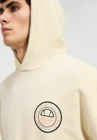 Sudadera beige con un logo redondo impreso que presenta contornos negros y un interior melocotón. Tejido suave, diseño casual con capucha y cordones.